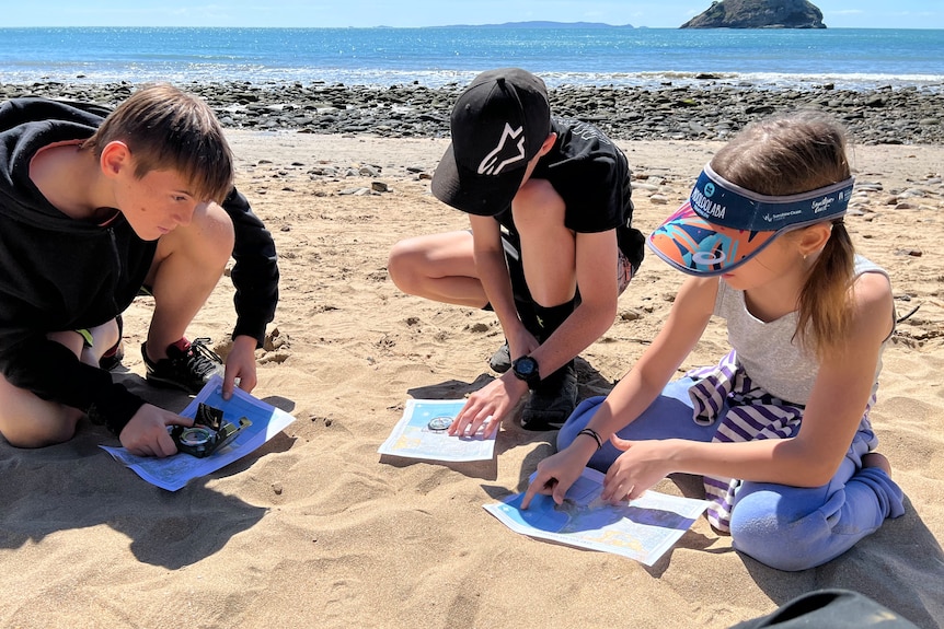 Trois jeunes enfants sur une plage regardant attentivement une carte.