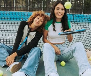 Deux filles en jean assises près d’un filet de tennis, souriantes, avec des balles de tennis