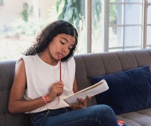 Adolescente assise sur un canapé, tenant un bloc-notes dans une main et un crayon porté à la bouche de l’autre