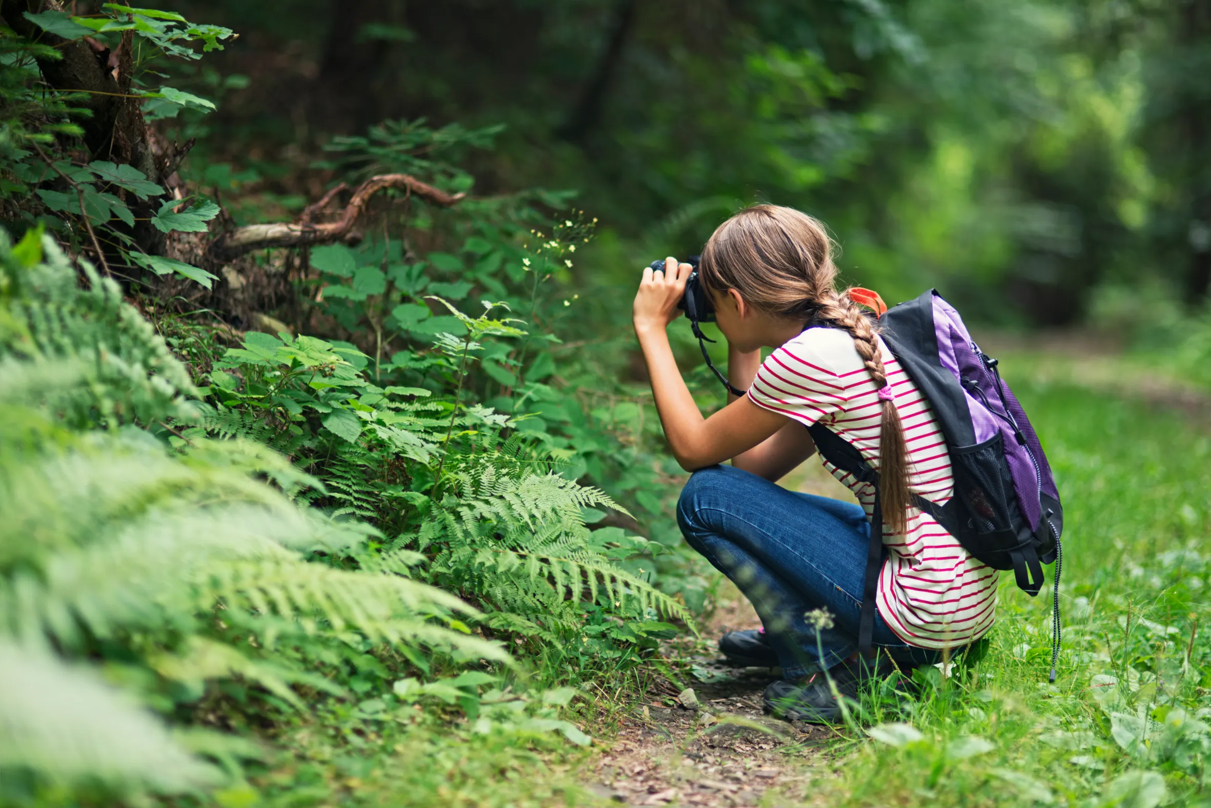 Jeune femme portant un sac à dos et tenant un appareil photo, accroupie dans une forêt.